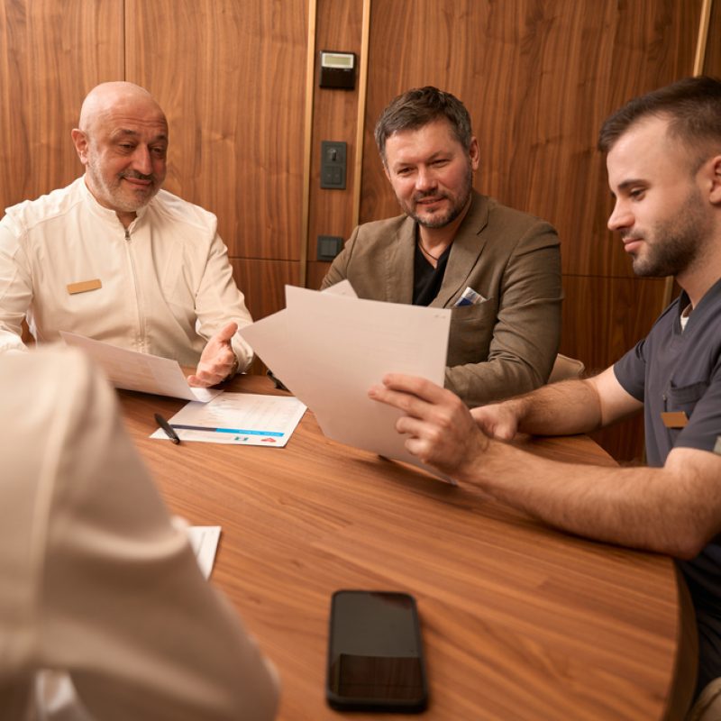 Smiling man sitting at table near young doctor and looking at prescription in the office